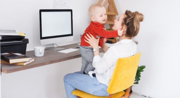 Side view of charming young stylish mother holding in hands and a cute baby sitting at the desk on white wall background. Place for advertising